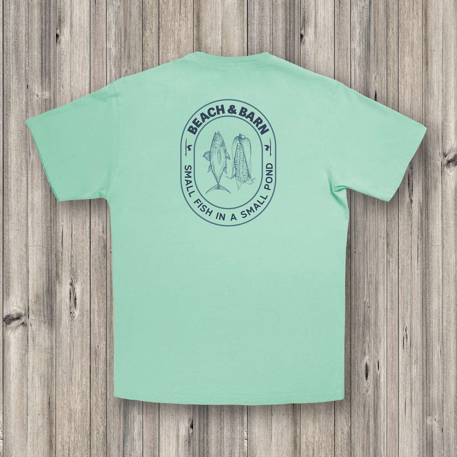 A light green T-shirt with a blue graphic on the back showing two fish and the text “BEACH & BARN” above “SMALL FISH IN A SMALL POND,” laid flat on a wooden surface.