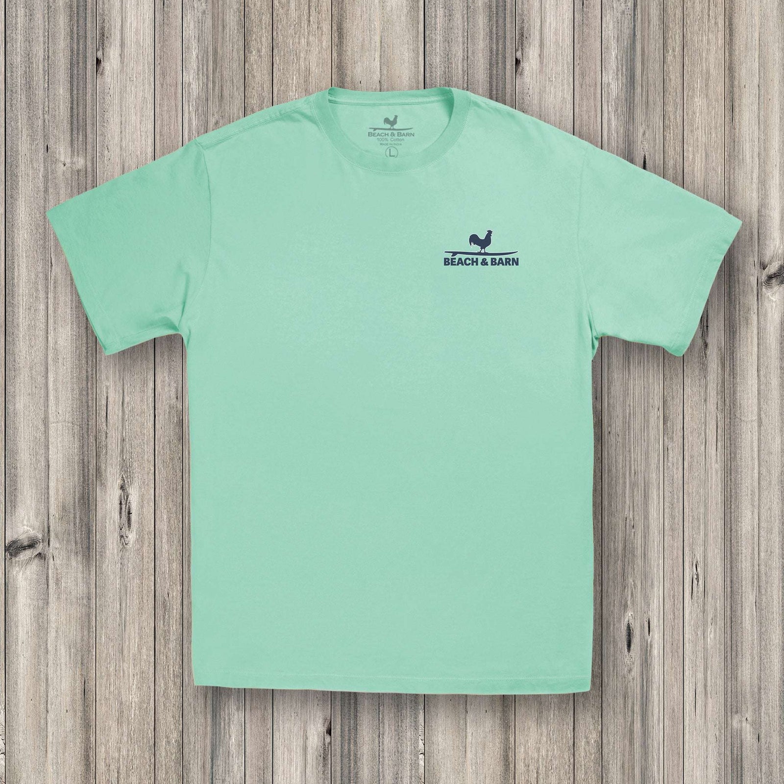 A light green T-shirt with a blue graphic on the back showing two fish and the text “BEACH & BARN” above “SMALL FISH IN A SMALL POND,” laid flat on a wooden surface.