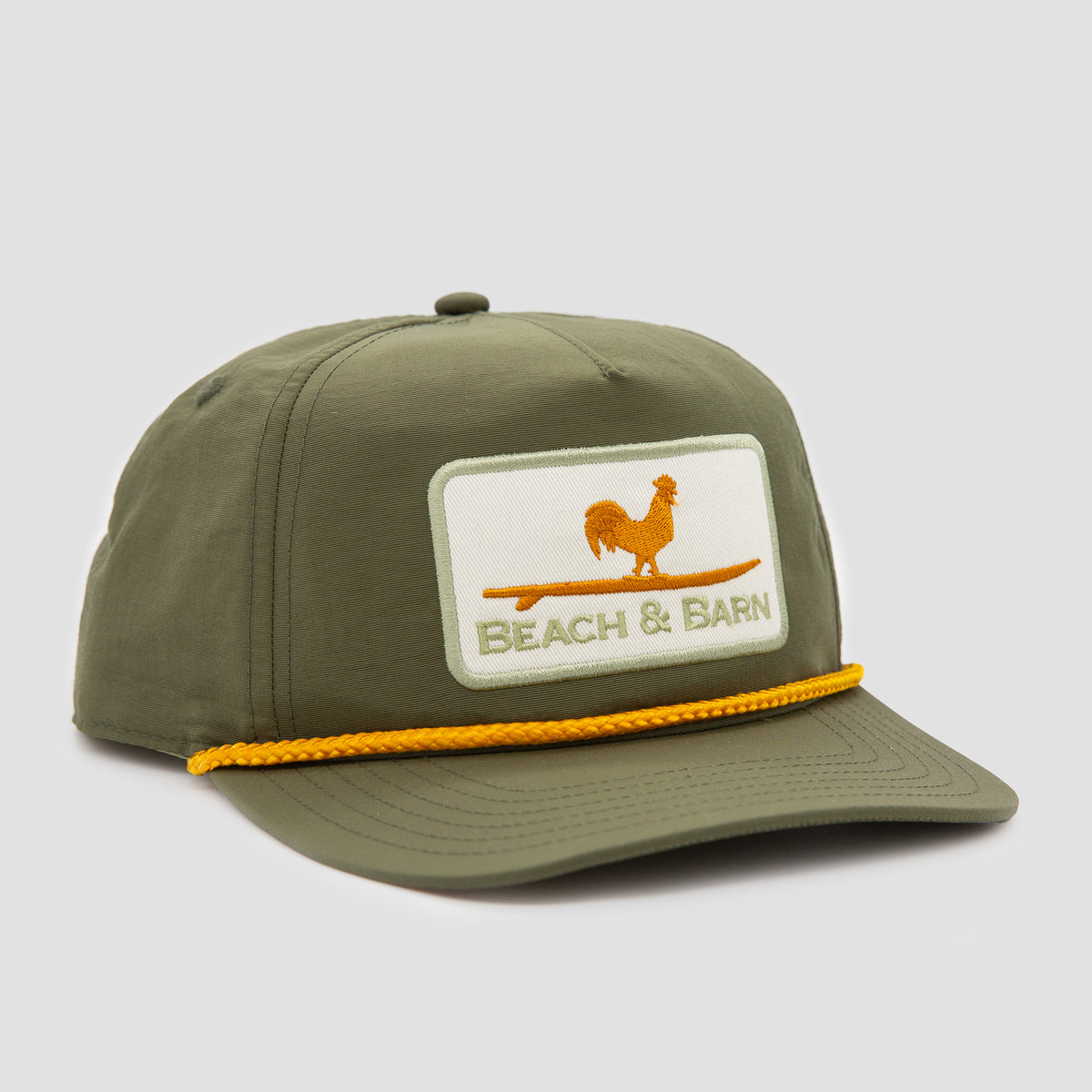 An olive green baseball cap with a yellow rope detail above the brim. The front patch features an orange rooster standing on a surfboard and the text BEACH & BARN.