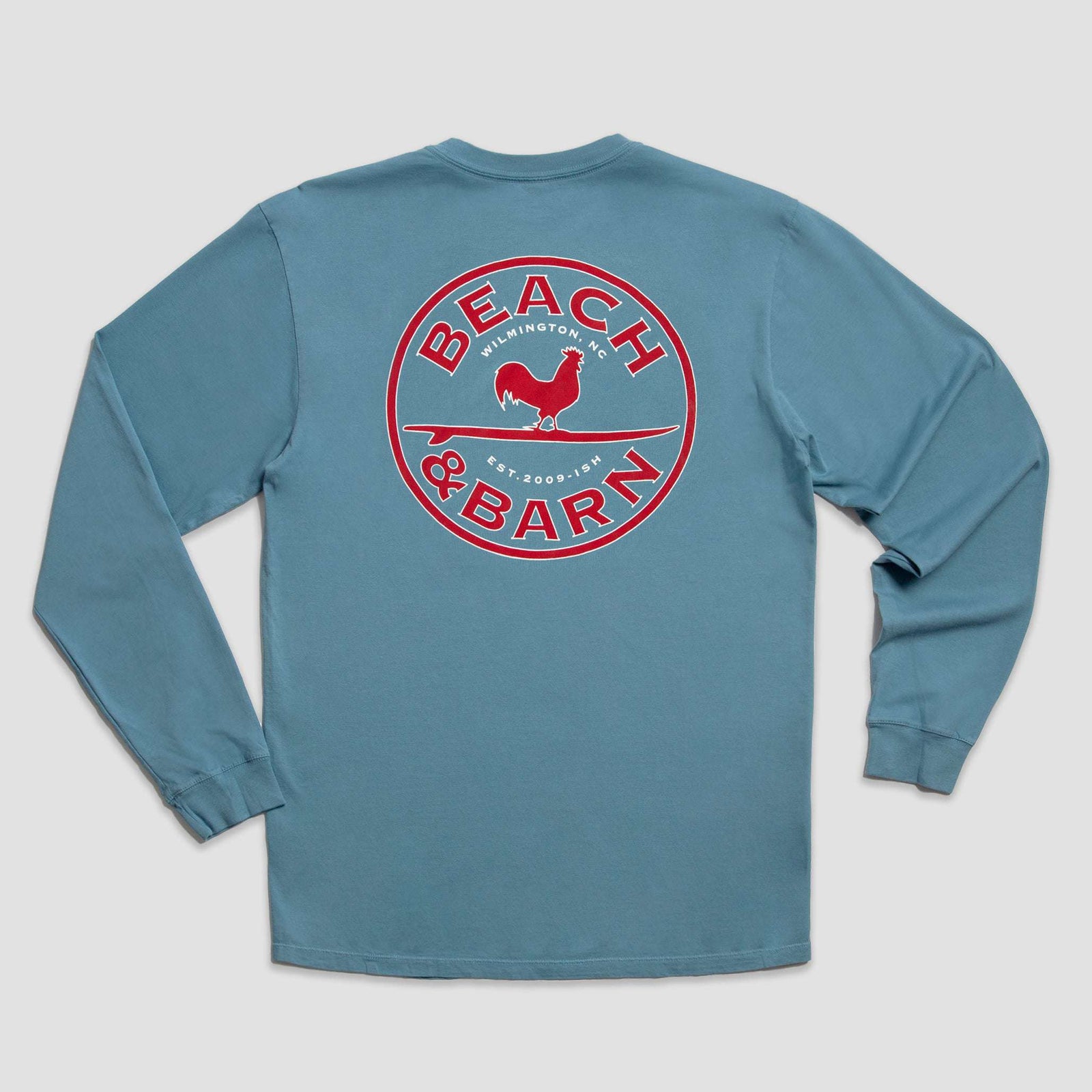 A blue long-sleeve shirt with a large red and white circular Beach & Barn logo on the back, featuring a rooster silhouette and the words Wilmington, NC, Est. 2016.